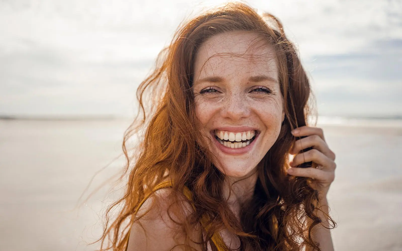 Smilende ung kvinne på stranden tar seg til håret med den ene hånden.