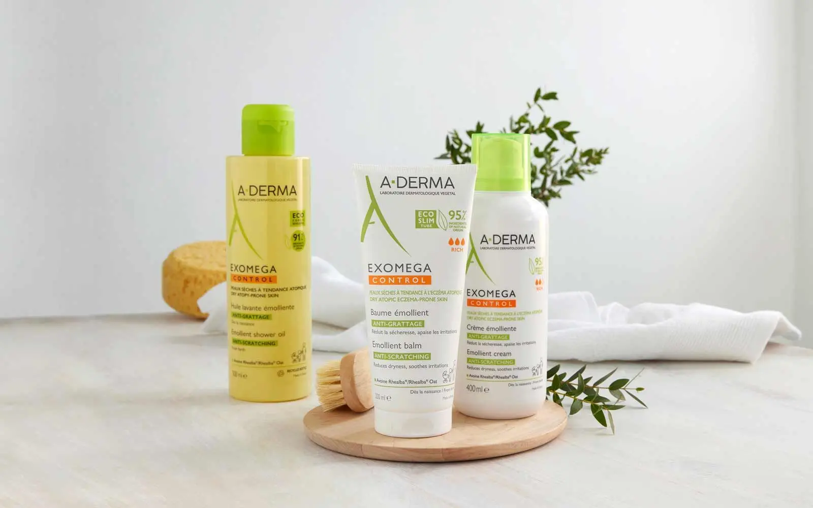 Diverse A-derma produkter står på et bord med blant annet grønne blader et håndkle og en svamp i bakgrunnen