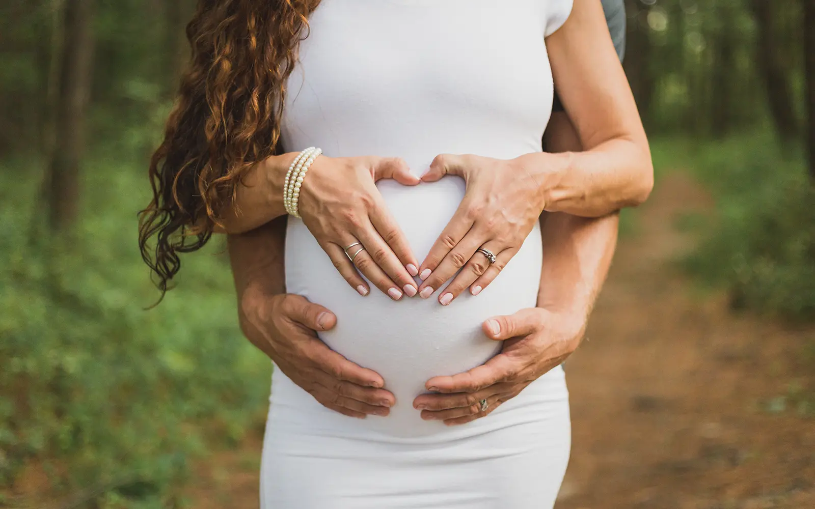 Gravid kvinne former hendene til et hjerte på magen, mens partner står bak henne og holder hendene under magen hennes