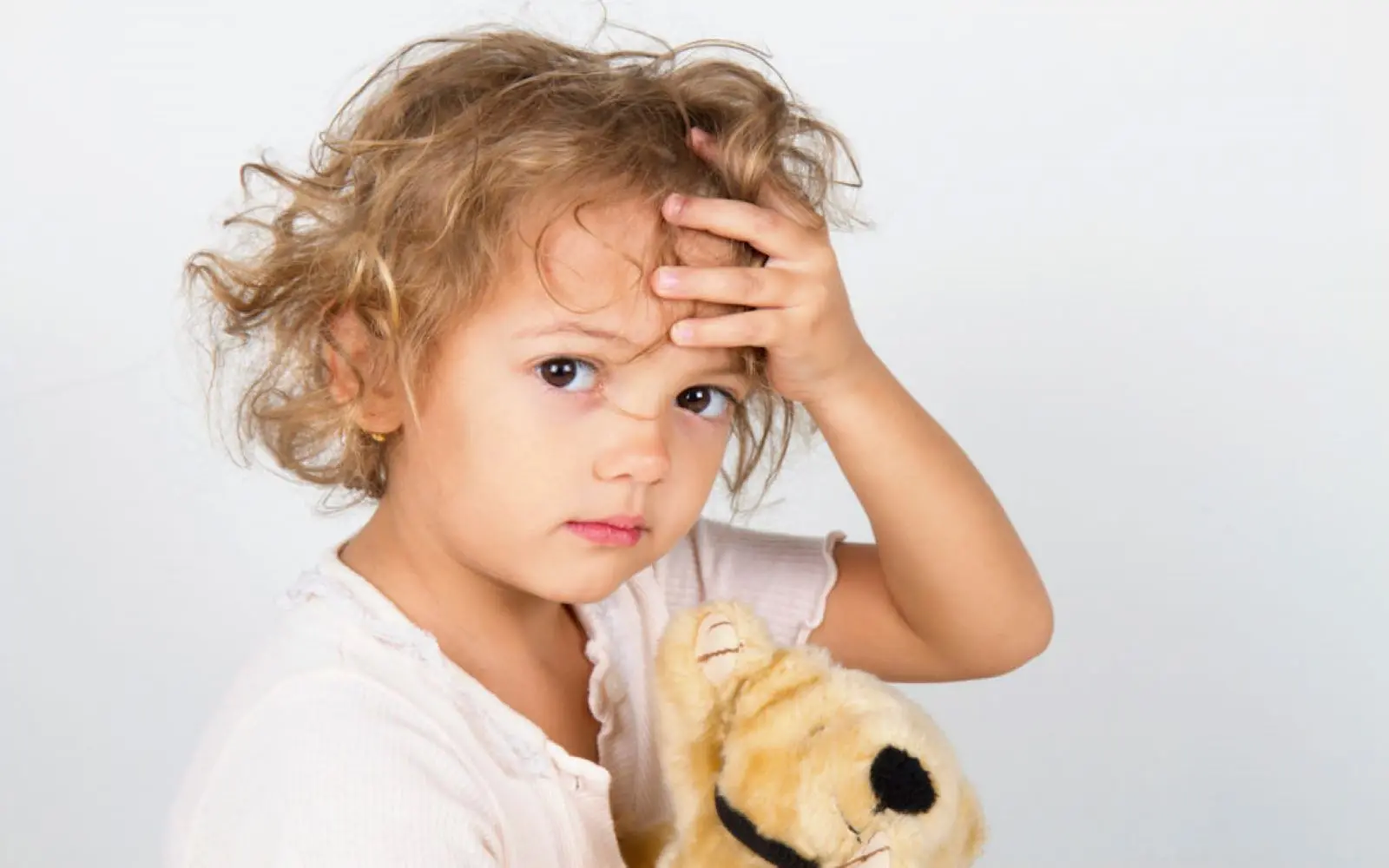 Et barn i 2-3 års alder ser inn i kameraet. Barnet holder en bamse i den ene hånden og tar seg til pannen med den andre.