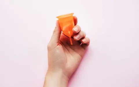En hånd holder en oransje menskopp opp-ned mot en lys rosa bakgrunn.