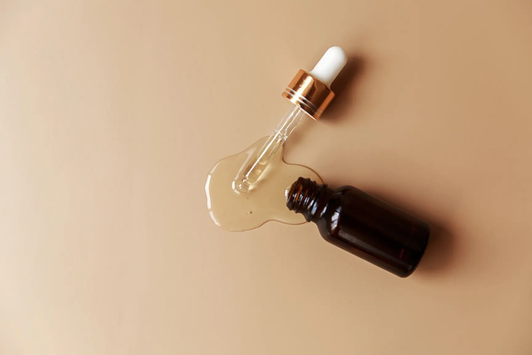 En brun glassflaske med hudpleieserum ligger åpen på en beige bakgrunn, med en pipette ved siden av og serum sølt utover under den.