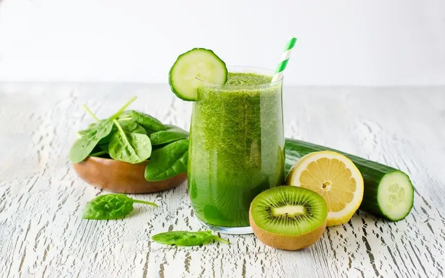 Et glass med grønn smoothie og sugerør. Agurk, sitron og kiwi ved siden av glasset.