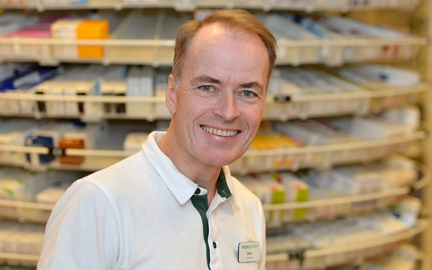 En man med hvit genser og VA navneskilt som ser mot kamera og smiler. I bakgrunnen hylle med medisiner.