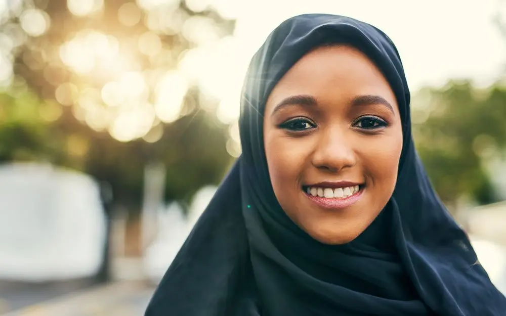 Ung kvinne med hijab smiler mot kamera i naturlig sollys. Bakgrunnen er mykt uskarp og varm i tonen.