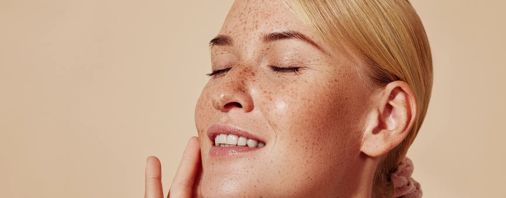 Nærbilde av en kvinne med fregner og glatt hud, med lukkede øyne og et avslappet smil, som forsiktig berører haken. Lys beige bakgrunn.