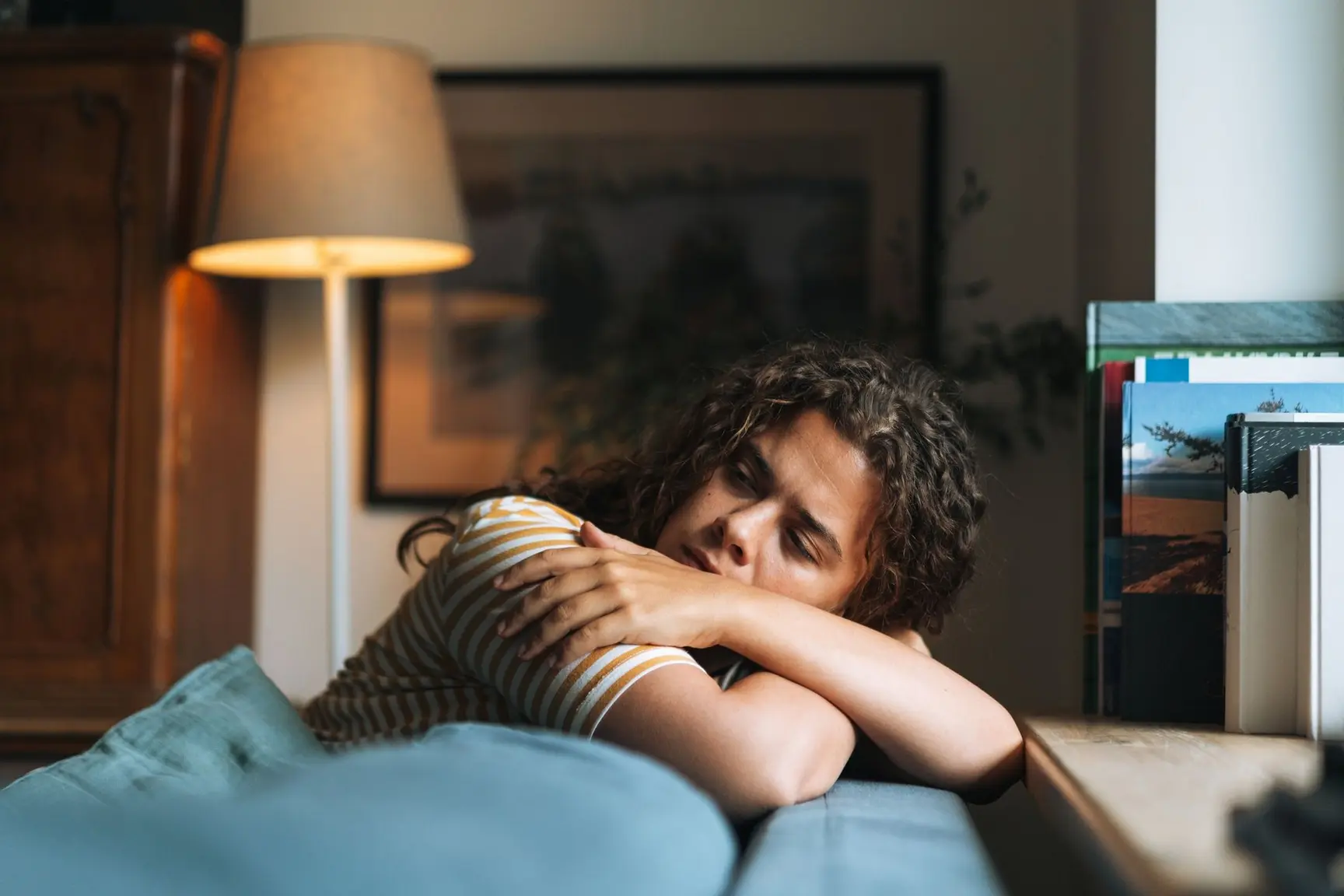 Kvinne sitter i sofaen med hodet hvilende på armene, og ser sliten og tankefull ut i et rolig stuerom.