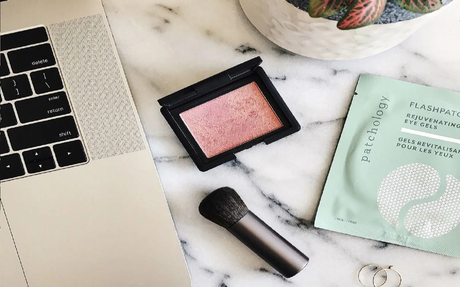 Flatlay-bilde av laptop på marmorbord sammen med blush, sminkekost, øyemasker og plante i bakgrunnen.