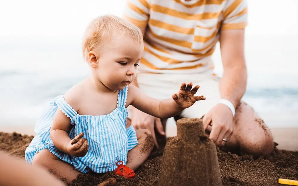 Et barn i hvit og blå stripete kjole leker med et sandslott på stranden mens en forelder sitter i bakgrunnen