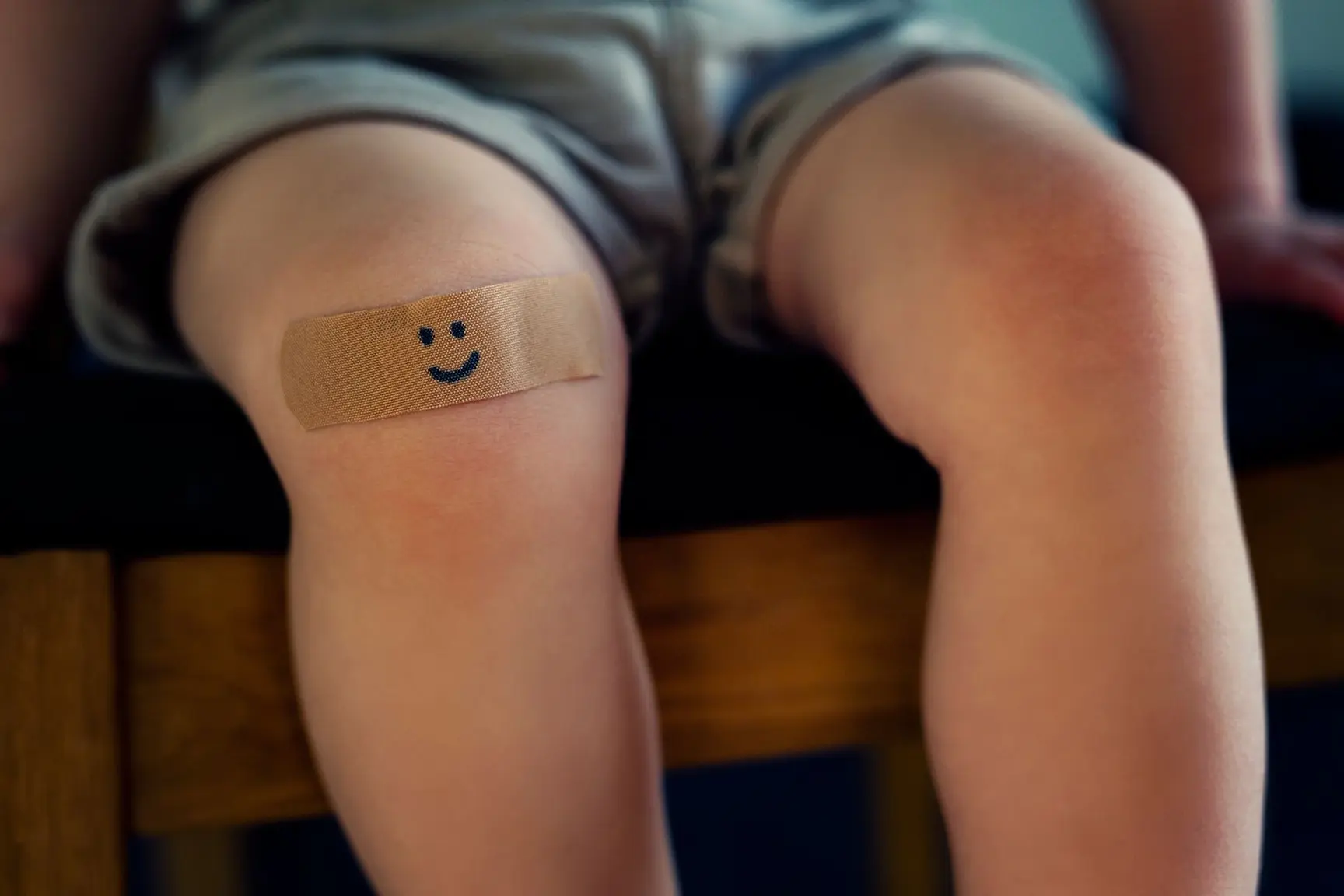 Et barn sitter på en stol med grønn shorts og har et plaster på kneet med et smilefjes på