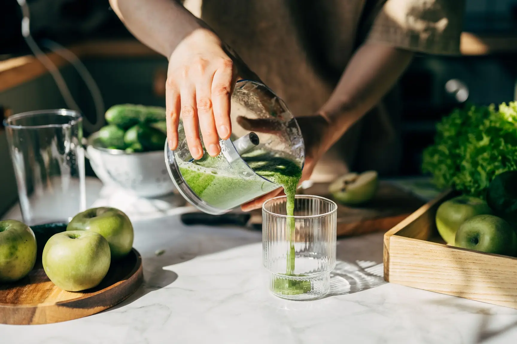 En person heller en grønn smoothie fra en blenderkanne over i et glass på en kjøkkenbenk omgitt av grønne epler, agurker og fersk salat i morgensol.