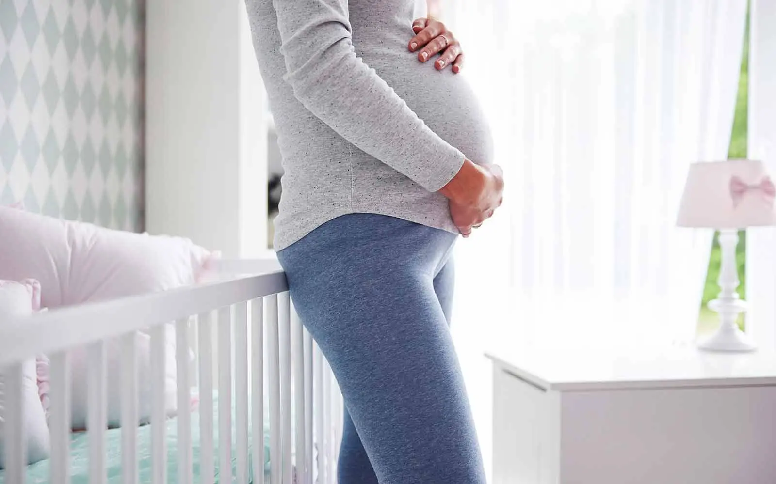 Bilde av en gravid dame som holder hendene over magen, stående ved en barneseng.