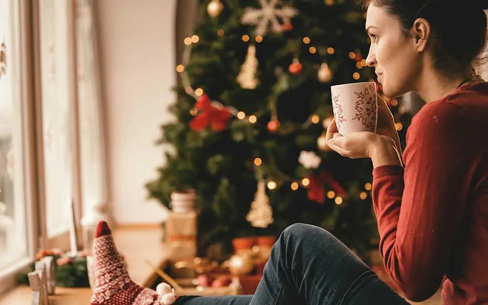 En dame som sitter på gulvet med en kopp mot ansiktet og et juletre i bakgrunnen