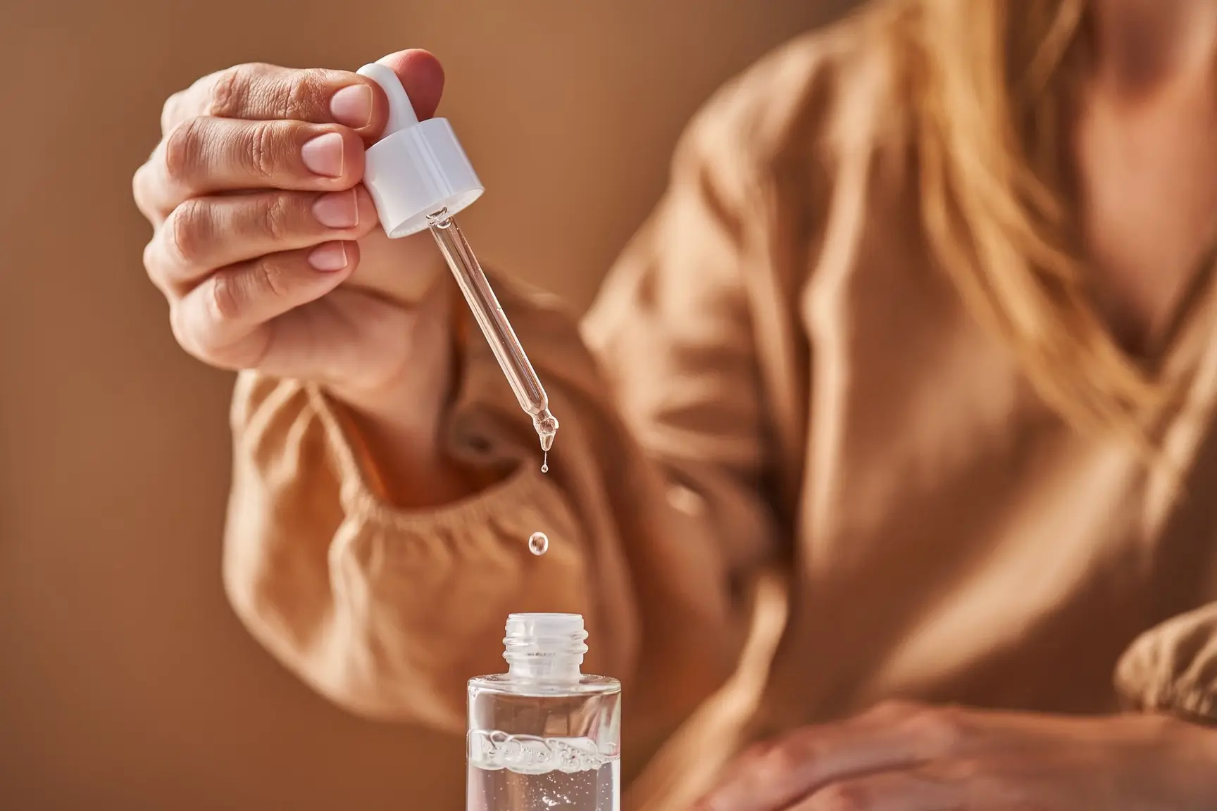Kvinnehånd holder pipette med hudpleieserum over en åpen flaske.