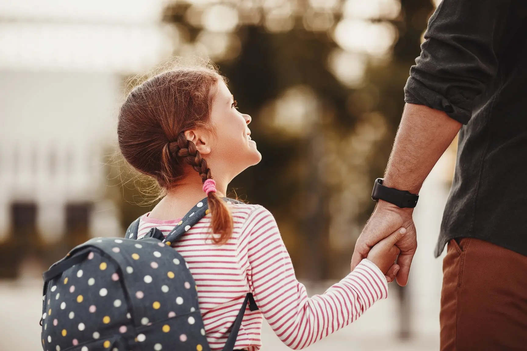 Bilde av en jente med skolesekk. Hun holder en voksen person i hånden, som hun smiler opp mot.