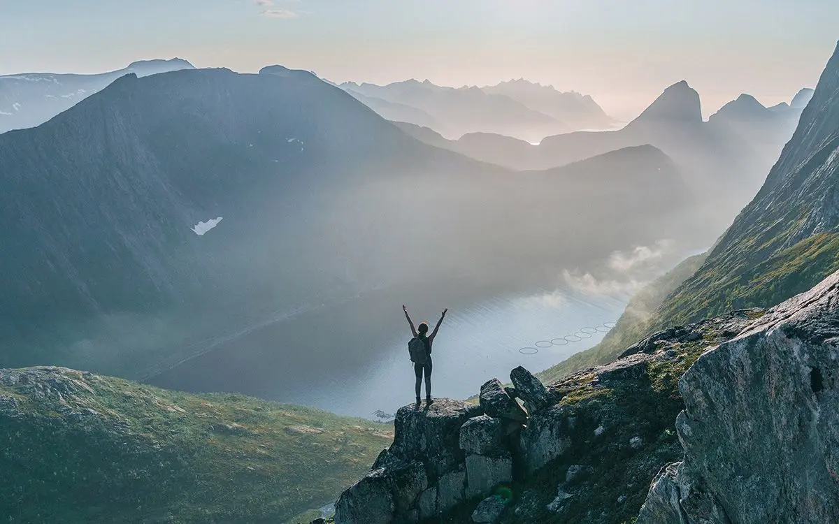 Et fjellandsskap og en person som står på en klippe med armene opp i været.
