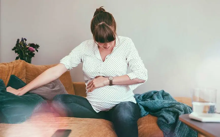 En gravid kvinne sitter på sofaen og holder seg på magen, trolig fordi riene har startet.