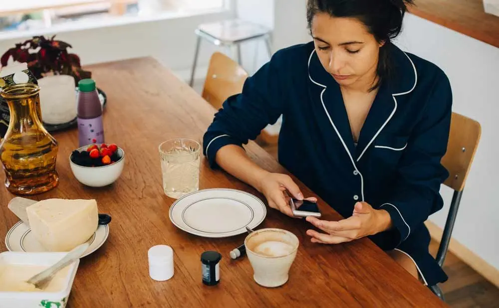En dame som sitter ved frokostbordet med mobilen i hånden. På bordet er det kaffe, ost,bær og medisinglass.
