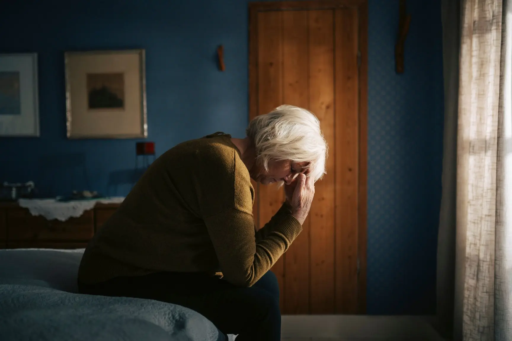 En eldre kvinne sitter på sengekanten med hodet i hånden, som om hun har vondt.
