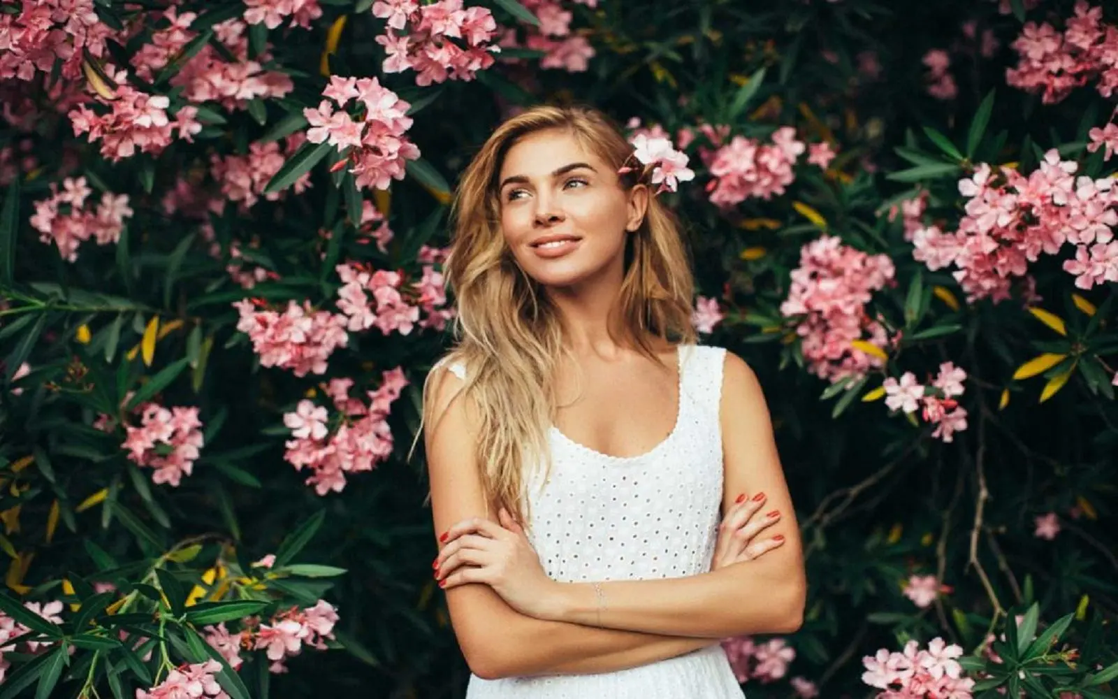 En dame med langt hår, hvit singlet og smiler utover med armene i kors. I bakgrunnen en grønn busk med rosa blomster.