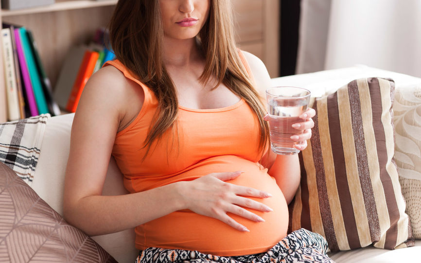 Bilde av en gravid kvinne sittende i en stol med et glass vann i hånden og andre hånd på magen.