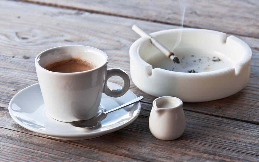 Nærbilde av en kopp med kaffe og et askebeger på et bord. På kanten av askebegeret hviler en tent sigarett.