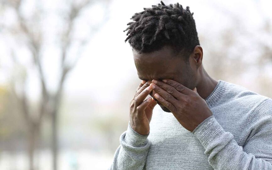 Bilde av ung mann som er utendørs. Bakgrunnen er uklar, men man kan skimte trær. Han presser begge hendene mot øynene.