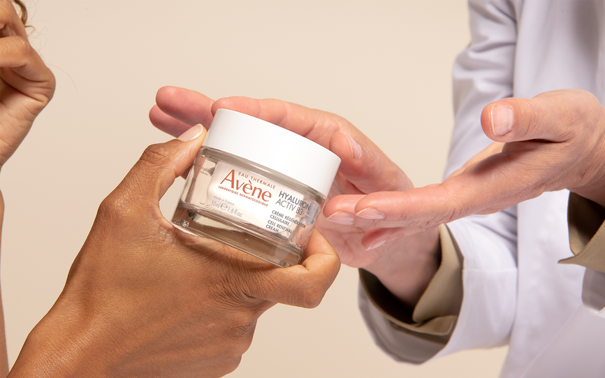 En arm ikledd laboratoriefrakk presenterer en krukke med Avene Active Hyaluron B3 ansiktskrem.