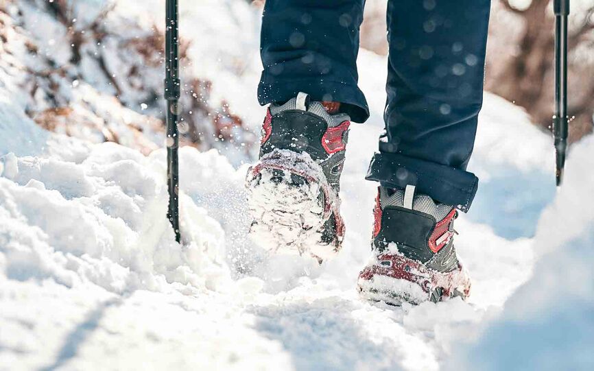 Bilde av person med vintersko som går i løs snø i skogsterreng. Bildet er tatt bakfra med fokus på skoene, som har brodder på.