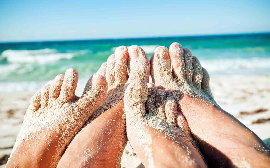 Nærbilde av føttene til et par som ligger på stranden. Føttene er delvis dekket med sand. I bakgrunnen er skyller bølger opp på stranden.