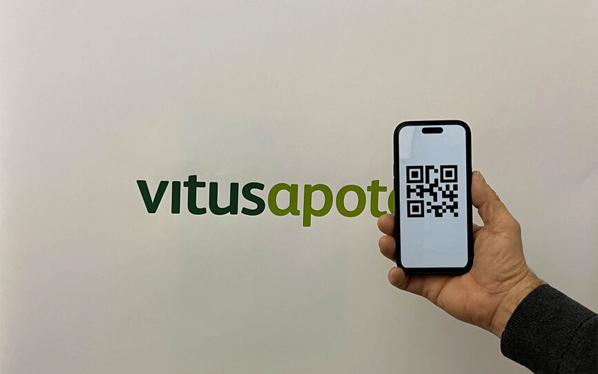 En hånd holder en mobiltelefon som har skannet en QR-kode. Vitusapoteklogo i bakgrunnen