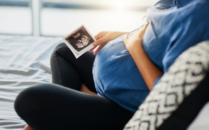 Nærbilde av en gravid kvinne som sitter i en seng. Hun holder en hånd på magen og i den andre hånden holder hun et ultralydbilde.
