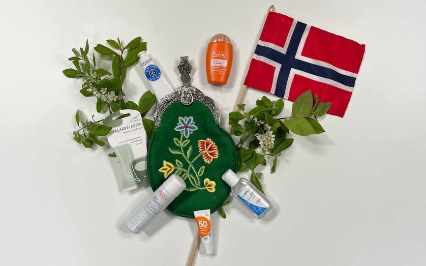 Bilde av en grønn bunadsveske omgitt av ulike produkter som solkrem, antibac, gnagsårplaster og kildevannspray. Et norsk flagg og bjørkekvister.