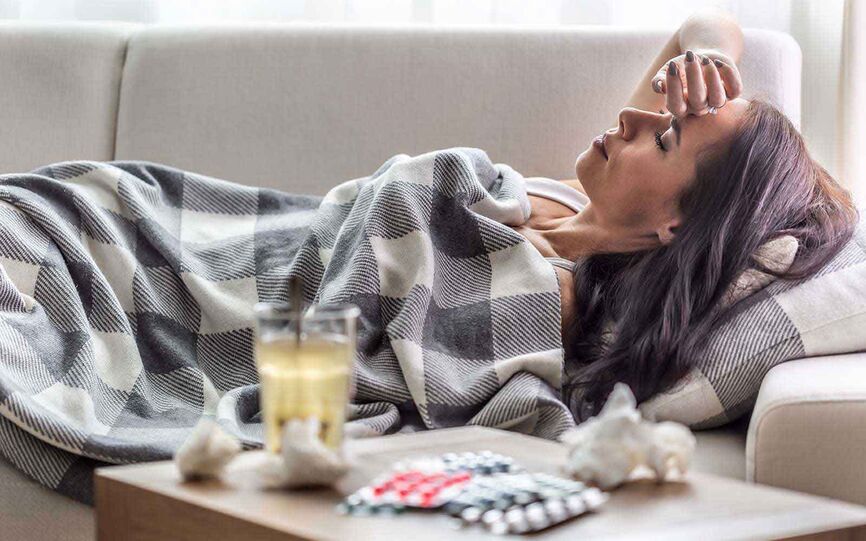Bilde av en kvinne som ligger på sofaen under et pledd. Hun lukker øynene og holder seg på pannen.