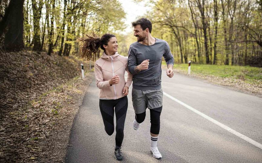 Bilde av smilende mann og kvinne som jogger sammen langs landevei omgitt av grønne trær.