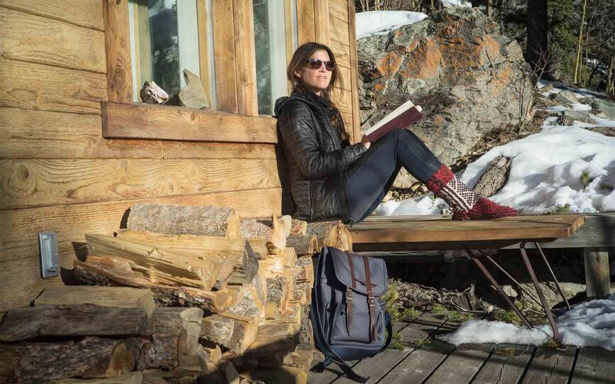 Bilde av en kvinne som sitter på en benk, lener seg mot en hyttevegg og leser en bok. Hun lener har solbriller på og i forkant står en tursekk.