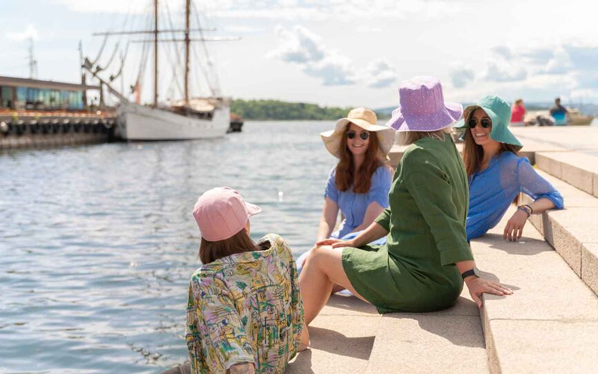 Bilde av fire smilende kvinner med solbriller og solhatter i ulike farger som sitter på en bryggekant. Det er lettskyet vær og solen reflekteres i sjøen.