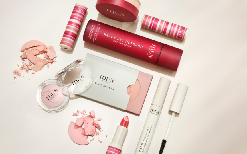 Et utvalg av makeup i duse nyanser fra IDUN minerals bestående av blush, leppestift, gloss og setting spray på creme-hvit bakgrunn.