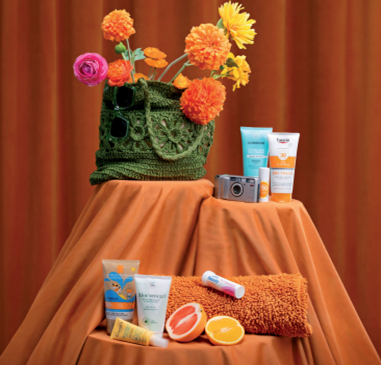 Bilde av en grønn standveske fyllt med blomster mot oransje bakgrunn. I forkant vises apotekvarer som kan være lurt å ha med på stranden.